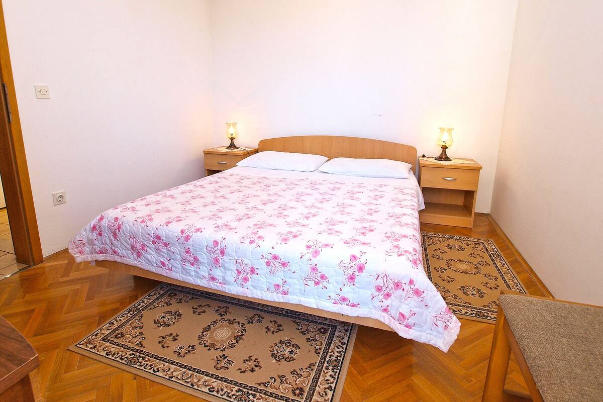 A bedroom with a wooden bed, two bedside tables with lamps, a patterned bedspread, and two rugs on the wooden floor.