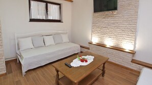 A room with a white sofa, a wooden table with a remote control and a lace doily, and a stone-textured wall with a TV mounted above. - Apartment 772 (Fazana)