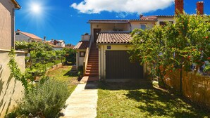 A residential property with a paved driveway leading to a front door, a small garden area with various plants, and a clear blue sky. - Apartment 785 (Pula)