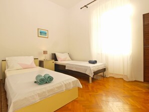A hotel room with two beds, a wooden floor, a window with curtains, and a small bedside table with a lamp. - Apartment 792 (Pula)