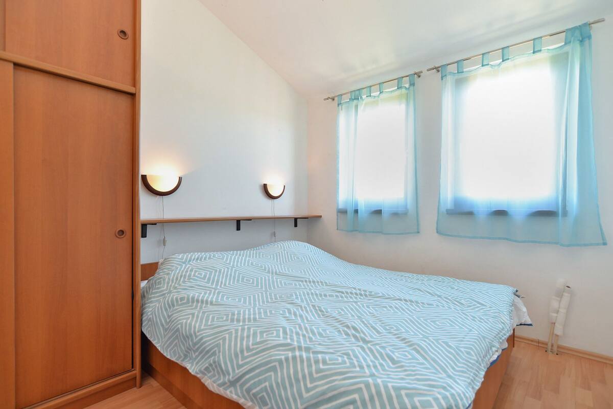 A bedroom with a bed, wooden wardrobe, two wall-mounted lights, and a window with sheer curtains.