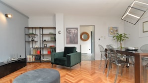 A modern living room with a green armchair, a round ottoman, a wooden herringbone floor, a bookshelf, and a dining area with clear chairs and a wooden table. - Apartment 843 (Fazana)
