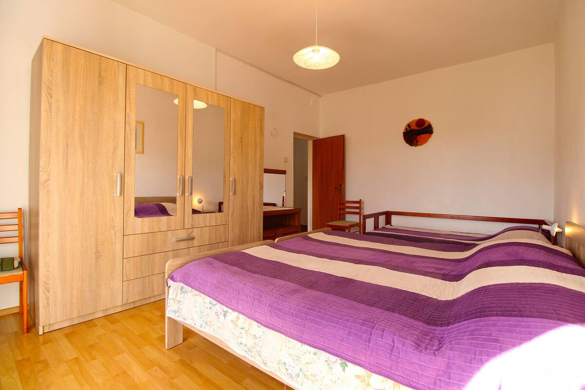 A bedroom with a large wardrobe, a bed with a purple and white striped bedspread, and a small desk with a chair.