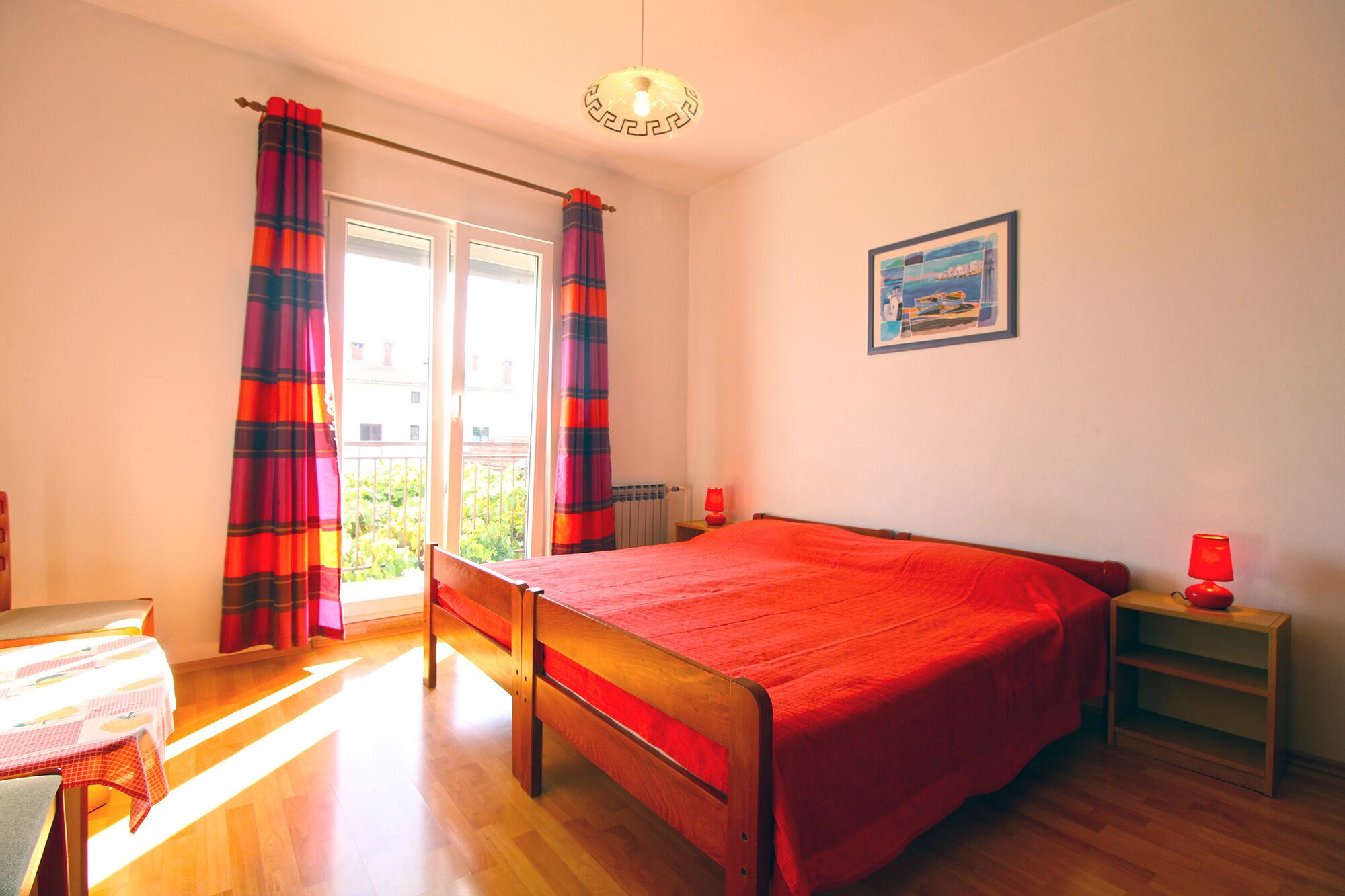 Una camera da letto con un letto, un tavolo, una sedia, una finestra con tende, un termosifone e un quadro appeso al muro.