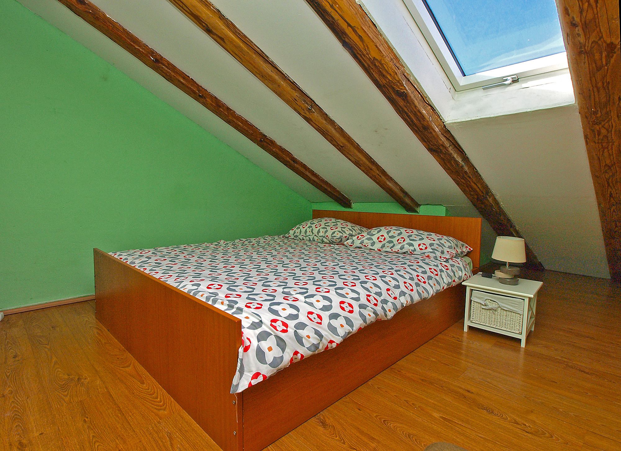 A bedroom with a sloped ceiling, a wooden bed frame, a patterned bedspread, a bedside table with a lamp, and a window with a view of the sky.