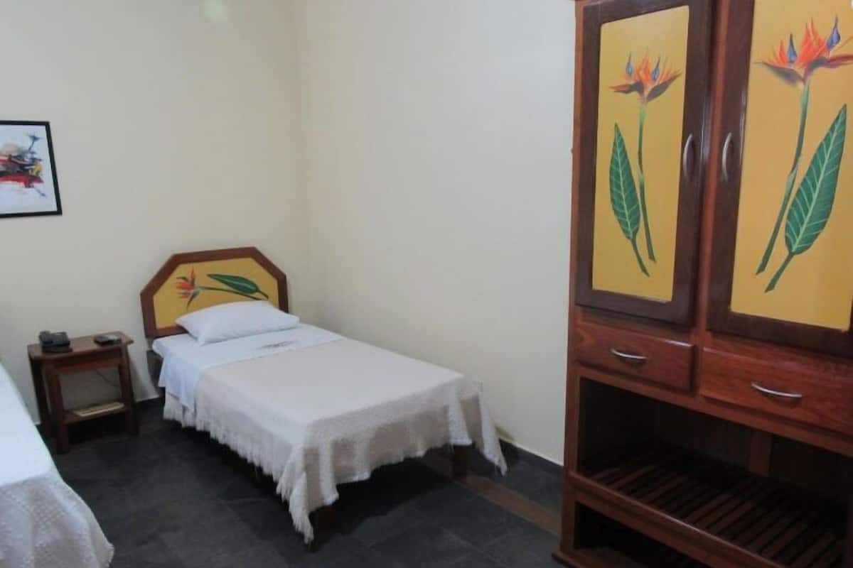 Um quarto pequeno e simples com uma cama, um criado-mudo e um guarda-roupa de madeira com motivos florais.