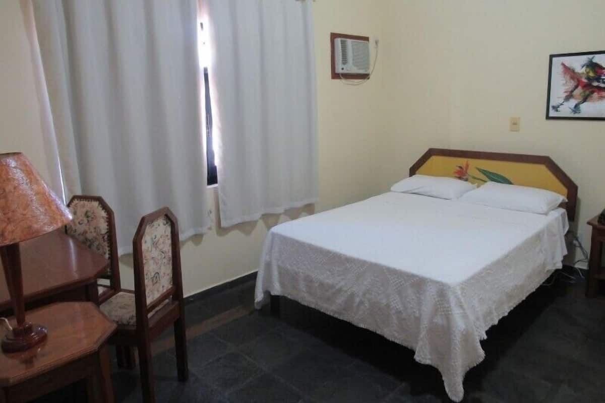 Uma cama de solteiro com cabeceira de madeira, uma escrivaninha com abajur e uma cadeira.