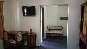 Een kamer met een aan de muur bevestigde televisie, een houten bank met gestreepte kussens, een klein tafeltje met een plant in pot, en een deur naar een andere kamer.