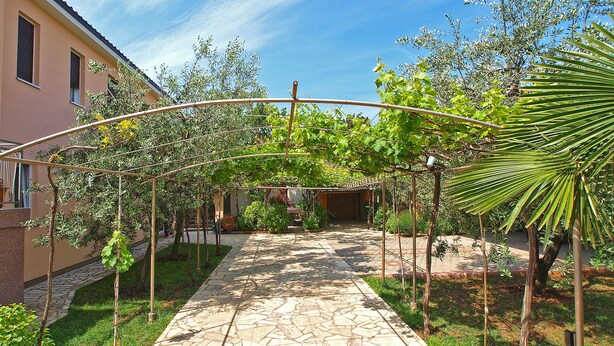 En stensti fører til et overdækket område med et pergola, omgivet af grøn vegetation og en bygning.