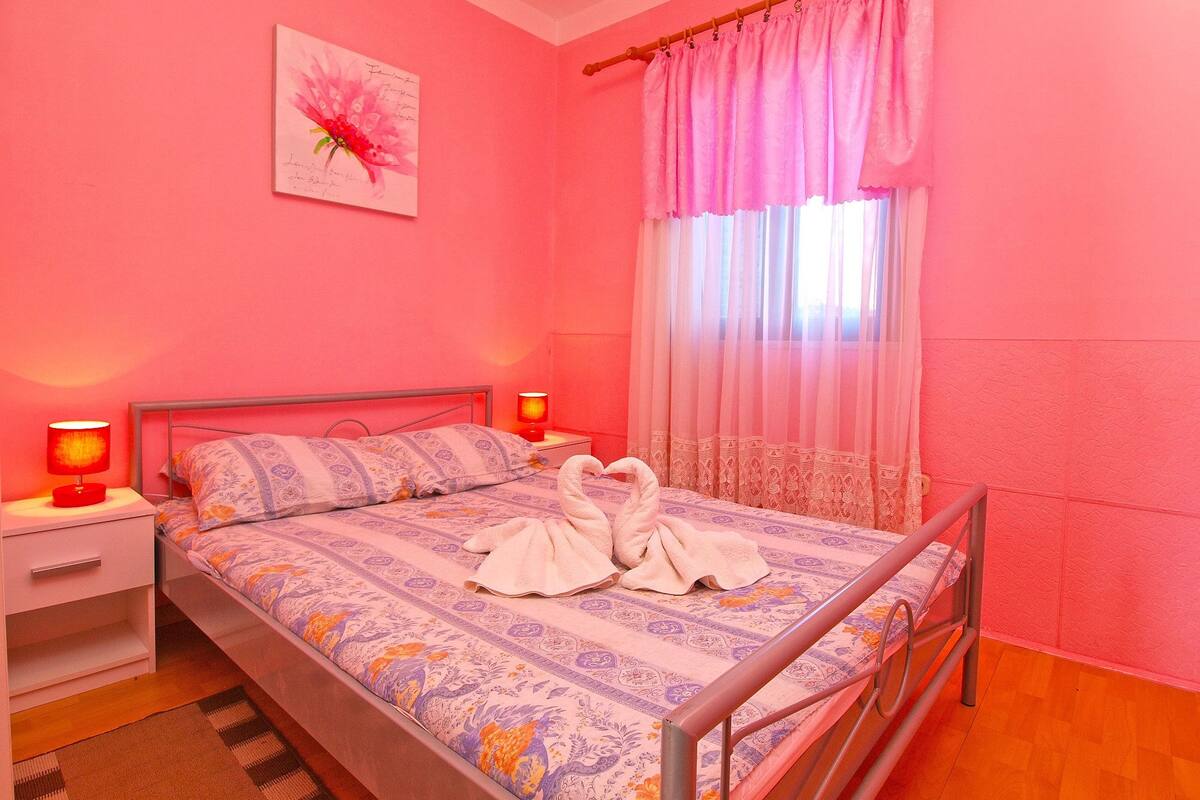 A pink-themed bedroom with a bed, two bedside lamps, a window with sheer curtains, and a floral artwork on the wall.