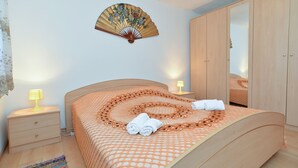 Un lit bien fait, recouvert d’une couvre-lit à motifs, deux lampes de chevet, un ventilateur mural et une armoire en bois.