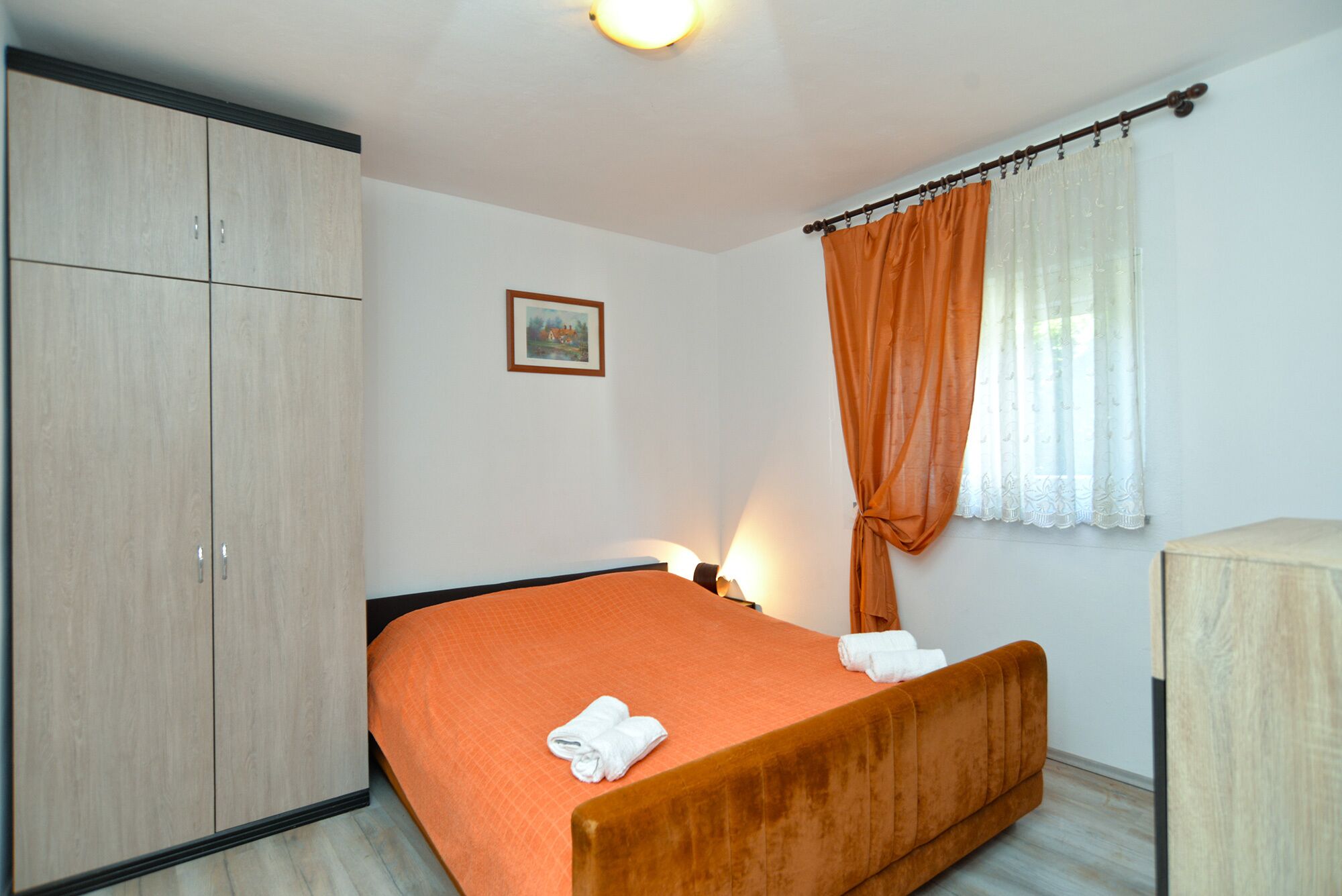 A bedroom with a bed, a wardrobe, a bedside table, a lamp, and a window with curtains.