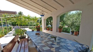 Eine Terrasse im Freien mit einem langen Tisch, der mit einer gemusterten Tischdecke bedeckt ist, Bänken, Blumentöpfen und einem hängenden Vogelkäfig.