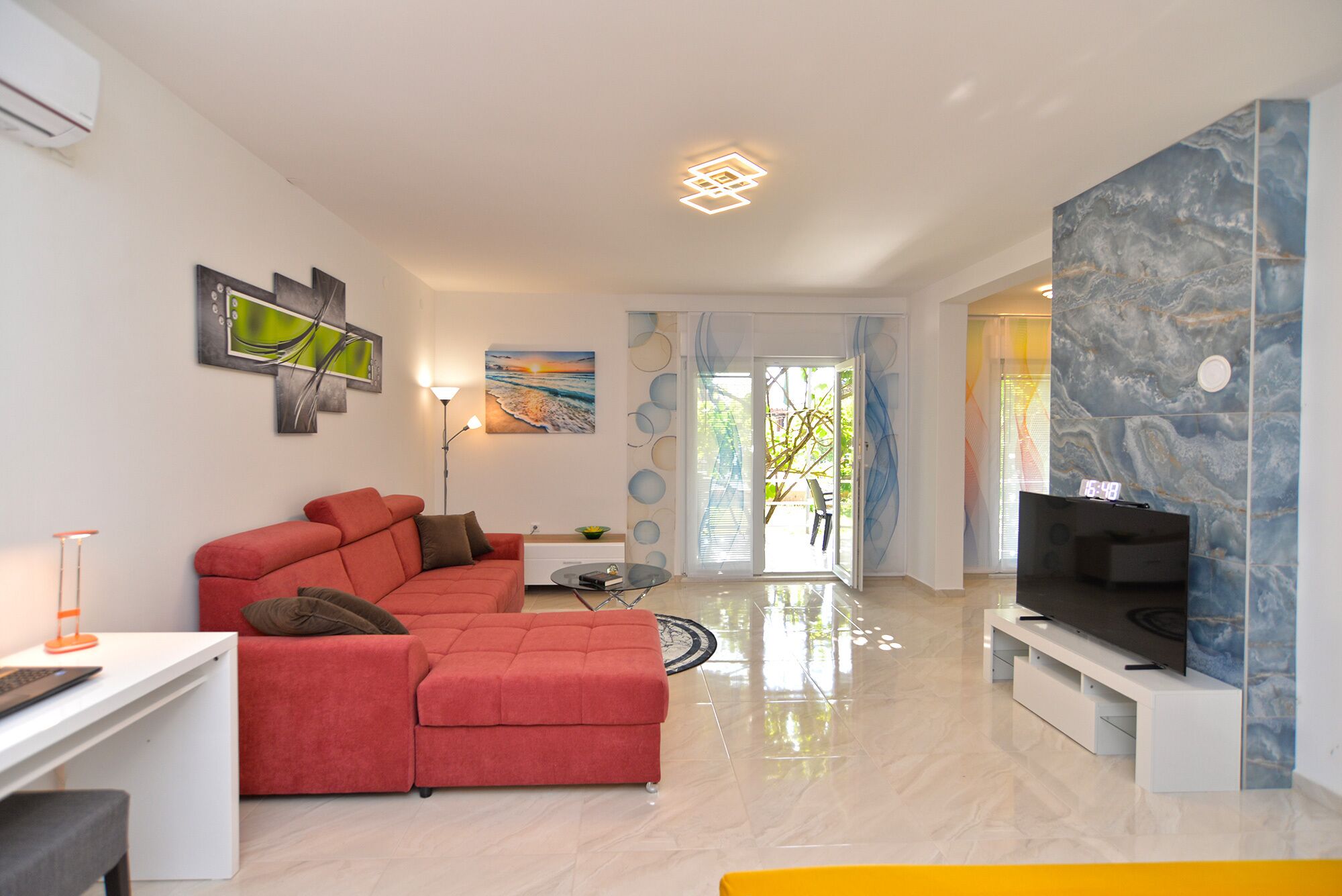 A modern living room with a red sectional sofa, a flat-screen TV on a white stand, and a marble-patterned wall.
