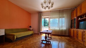 Ein Zimmer mit Fischgrätparkett, einem grünen Sofa, einem kleinen Tisch mit Spitzenbettdecke, einer Chandelieber und Holzschränken mit Glastüren.