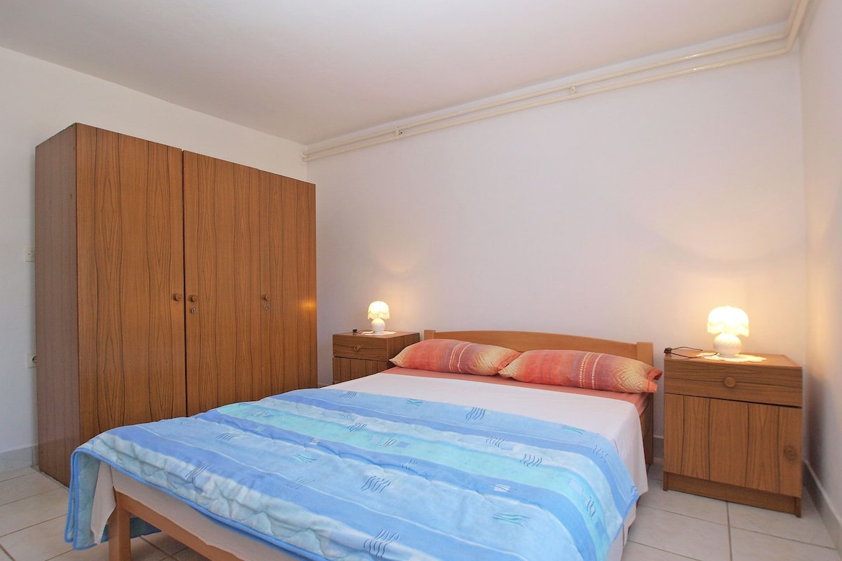 A bedroom with a bed, wooden wardrobe, bedside tables with lamps, and a tiled floor.