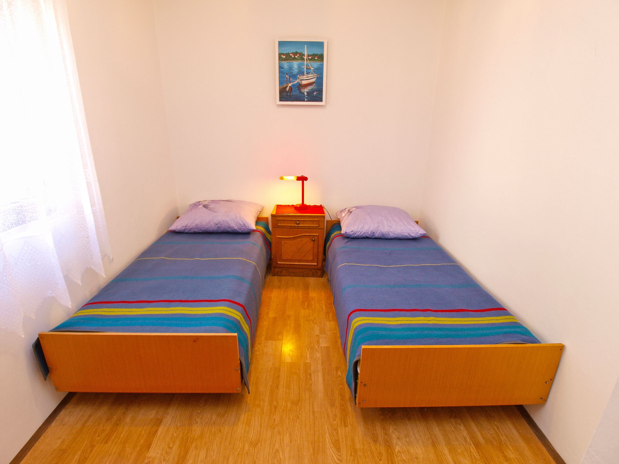 Una habitación pequeña y bien iluminada con dos camas individuales, piso de madera, una mesita de noche con lámpara y un cuadro en la pared.