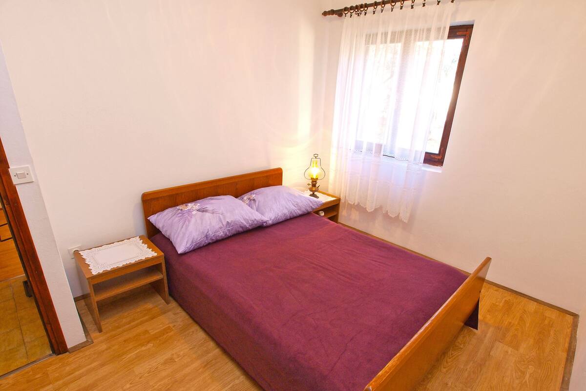 Un dormitorio con cama de madera, ropa de cama púrpura, una mesita de noche con lámpara y una ventana con cortinas translúcidas.