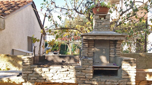 A stone-built outdoor structure with a chimney, possibly a barbecue area, surrounded by a garden with various plants and a building in the background. - Apartment 940 (Medulin)
