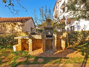 Cozinha externa de pedra com chaminé, rodeada por vegetação e um edifício branco ao fundo.