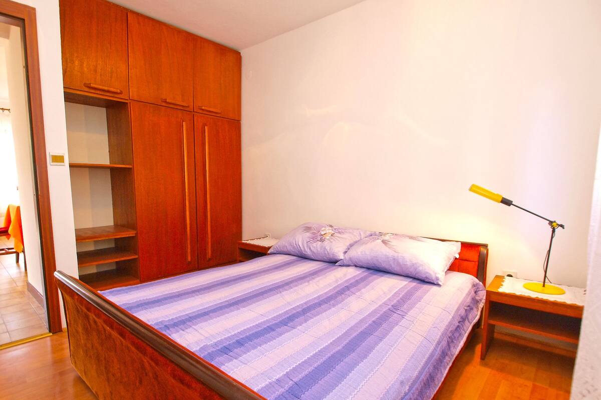 Un dormitorio con estructura de cama de madera, cubrecama a rayas, mesita de noche con lámpara y armario de madera.