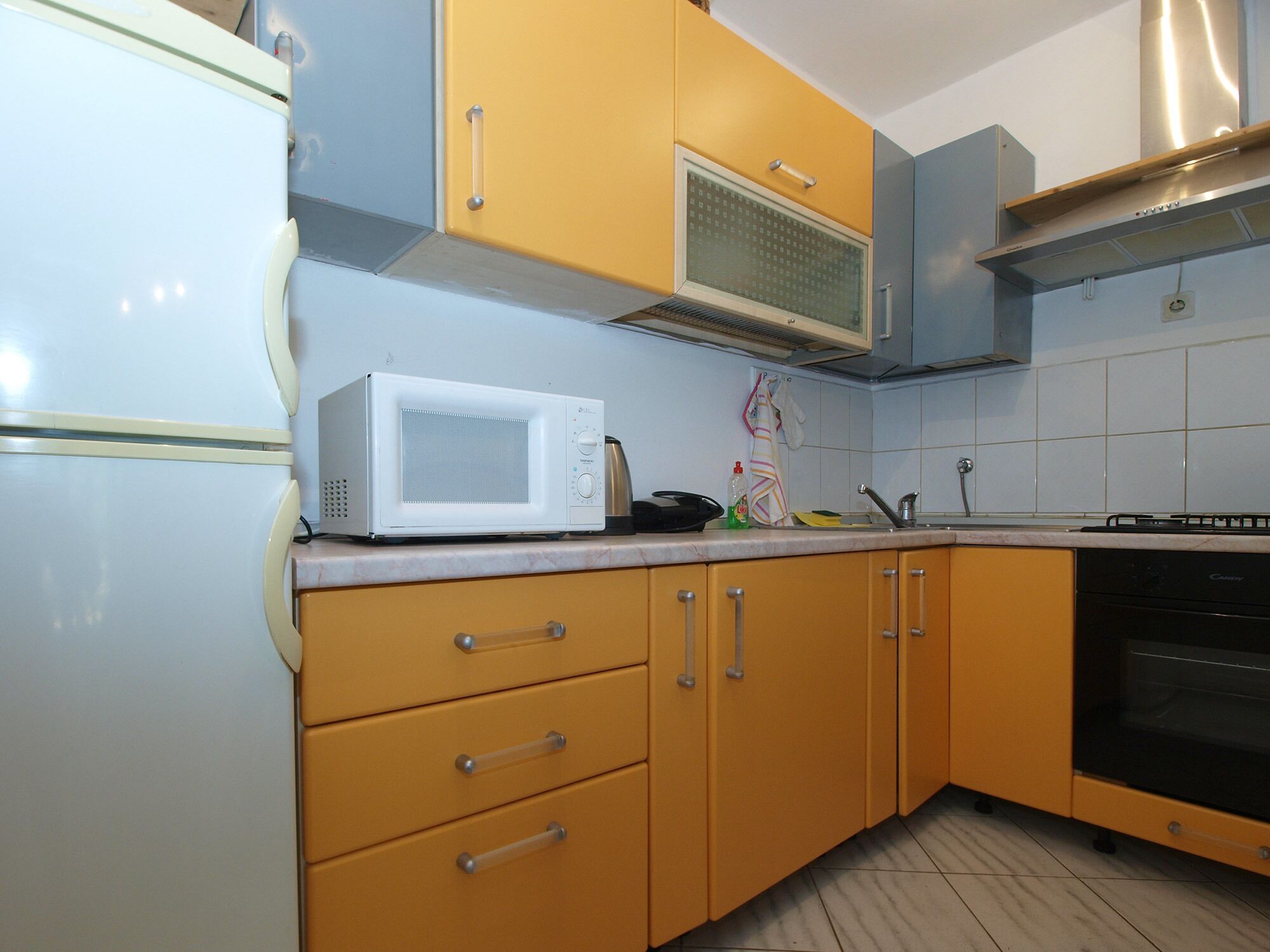 Una cocina con refrigerador blanco, microondas y estufa. Se observan gabinetes amarillos y un salpicadero de azulejos.