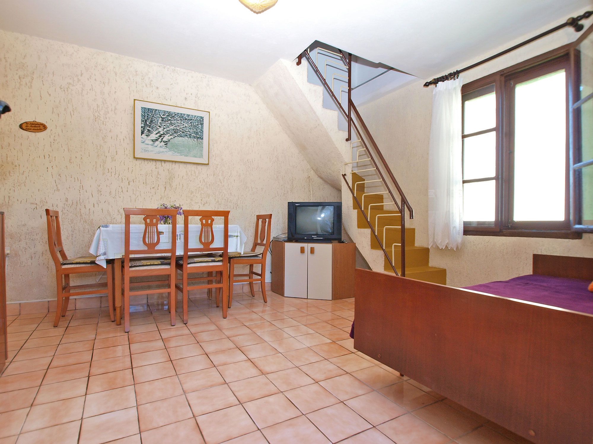 Un espace repas comprenant une table et des chaises, un téléviseur, un escalier et une fenêtre avec des rideaux.