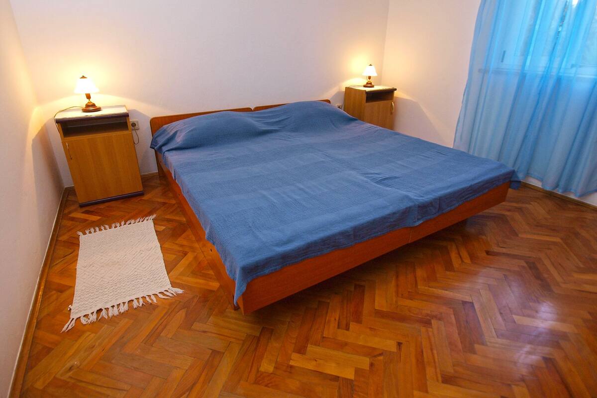 A bedroom with a wooden bed, two bedside tables with lamps, a white throw rug, and a window with blue curtains.