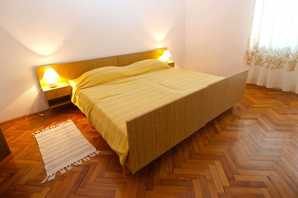 A single-bed room with a wooden headboard, two bedside lamps, a patterned rug, and a herringbone wooden floor.
