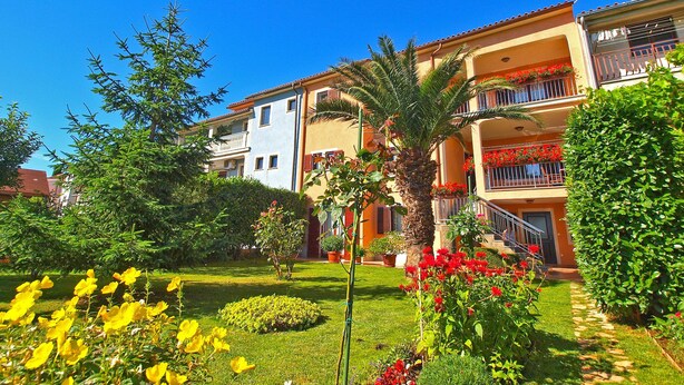 A multi-story building with balconies, a well-maintained garden with various plants and flowers, and a clear blue sky. - Apartment 979 (Medulin)
