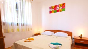 A neatly made bed with white linens, two bedside tables with lamps, a colorful wall art piece, and a window with sheer curtains. - Apartment 990 (Fazana)