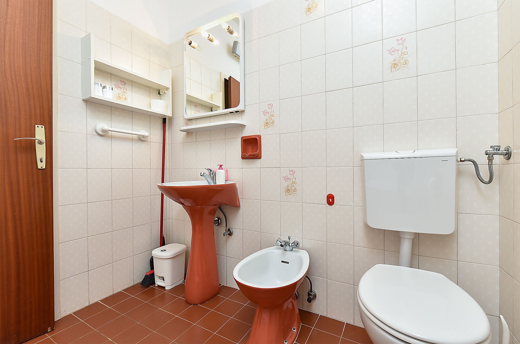 A bathroom with a pedestal sink, a bidet, a toilet, a mirror, and a shelf with toiletries.