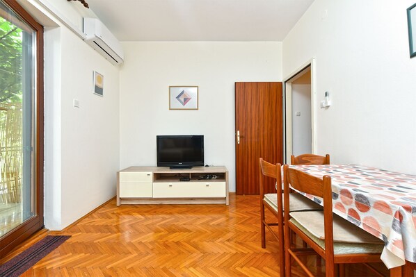 A small, well-lit room with a wooden herringbone floor, a television on a white cabinet, a wooden door, and a table with a patterned tablecloth.