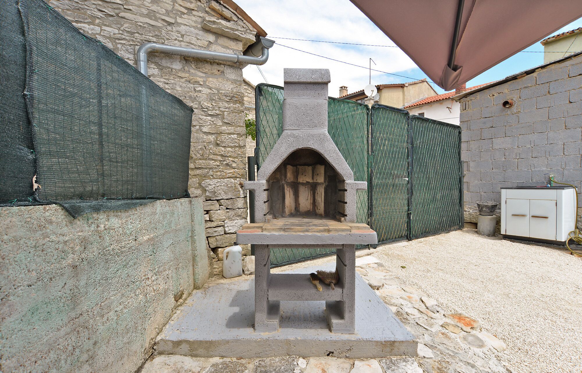 A stone-built outdoor oven with a chimney, surrounded by stone walls and a green fence.
