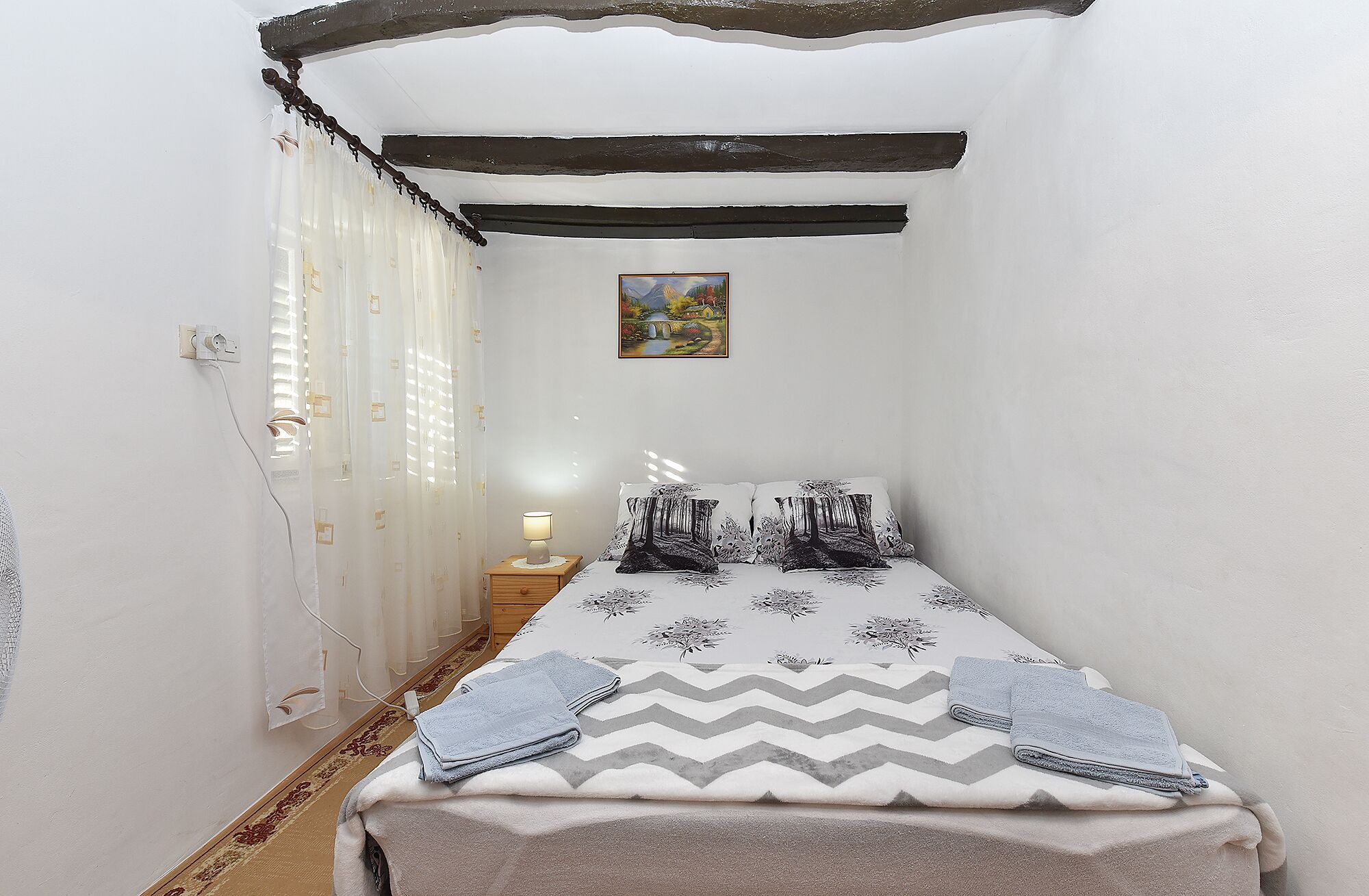 A small, cozy bedroom with a bed, a nightstand, a lamp, and a painting on the wall.