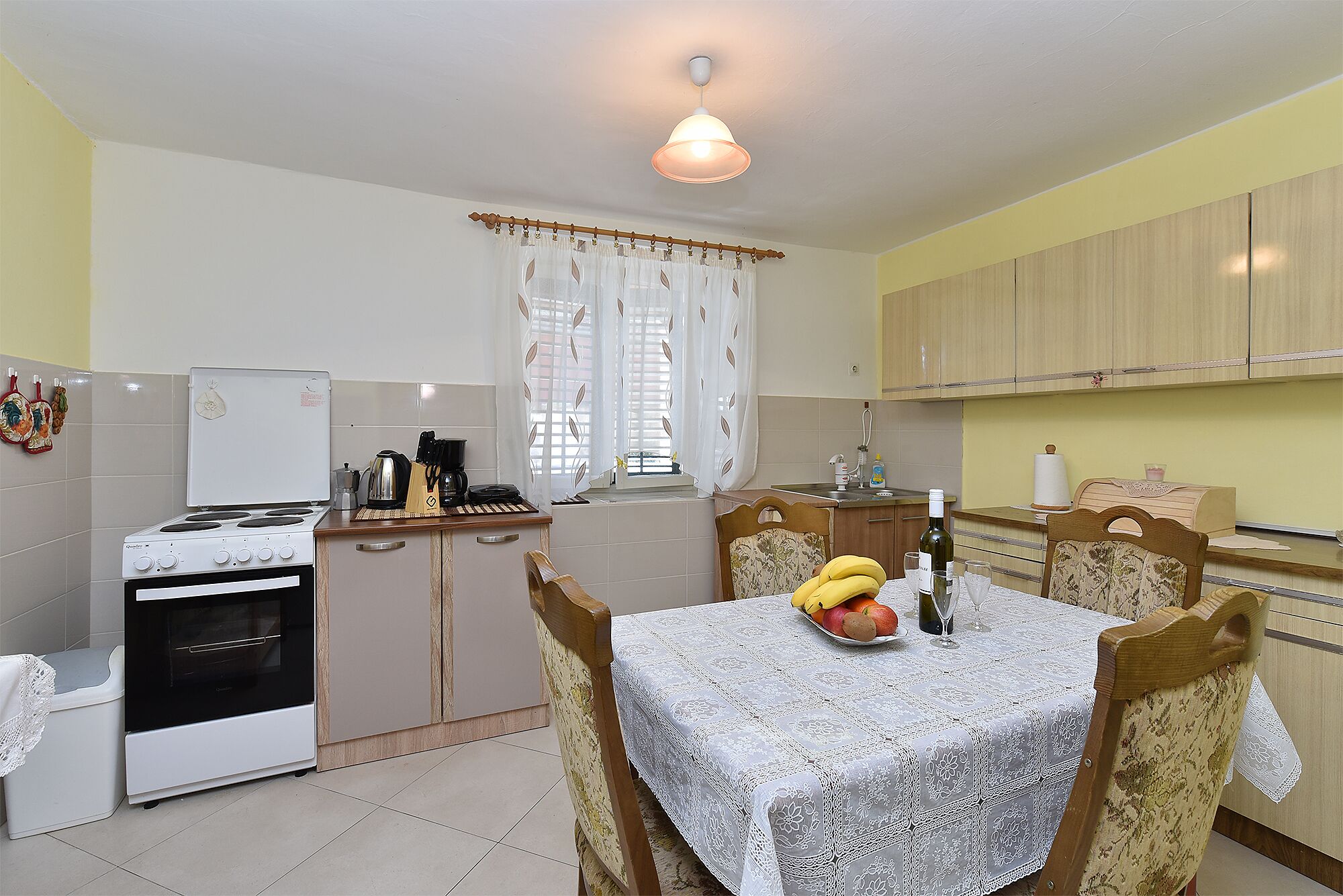 A compact kitchen with a stove, refrigerator, and wooden cabinets. There's a dining table with a lace tablecloth and a fruit bowl.