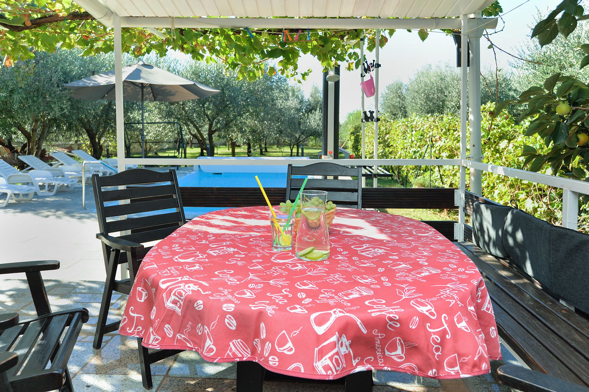Een overdekt terras met een tafel gedekt voor twee personen, met een rode tafelkleed met witte koffiekopmotieven.