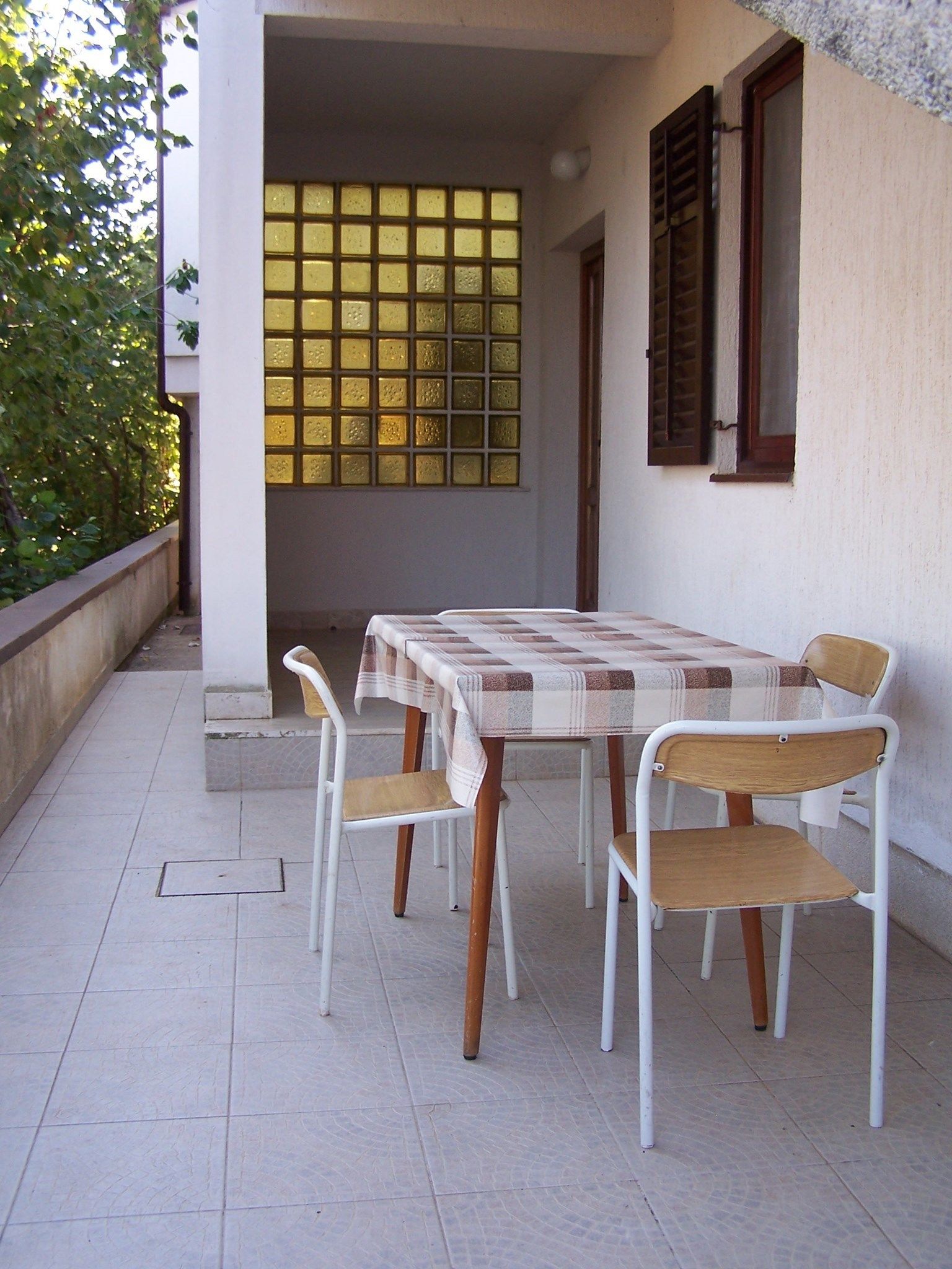 An outdoor patio with a table and chairs, a window with shutters, and a tiled floor.