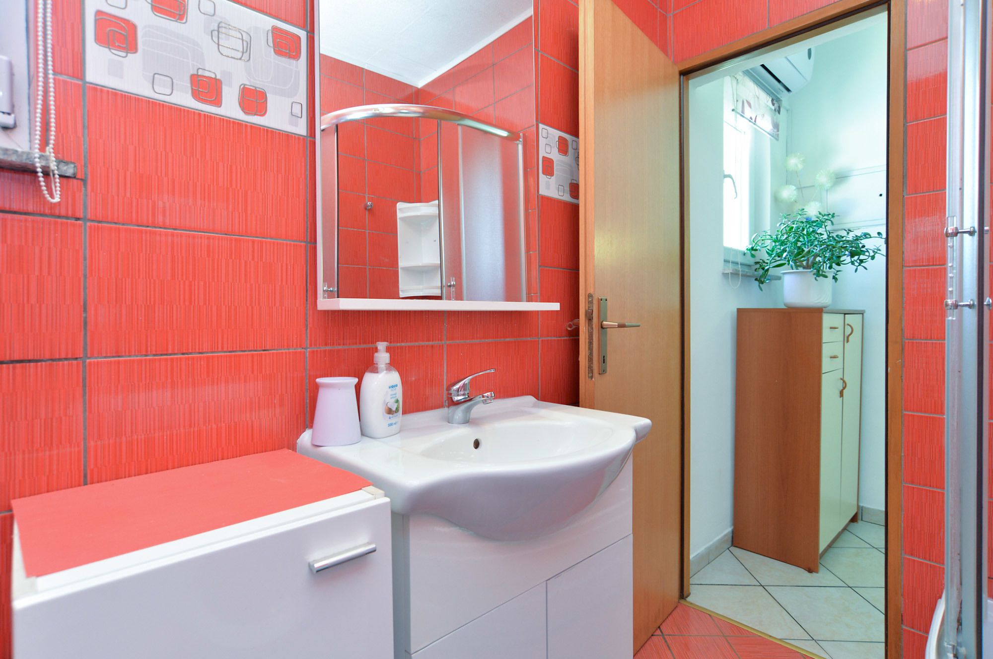 A bathroom with red tiled walls, a white sink, and a mirror. There is a door leading to another room with a plant.