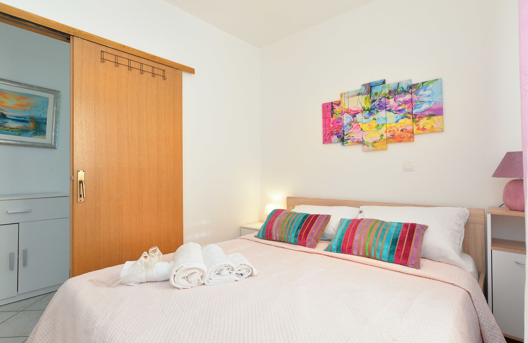 Un lit bien fait, avec du linge de lit blanc et des coussins rayés aux couleurs vives, une armoire en bois et un tableau accroché au mur.