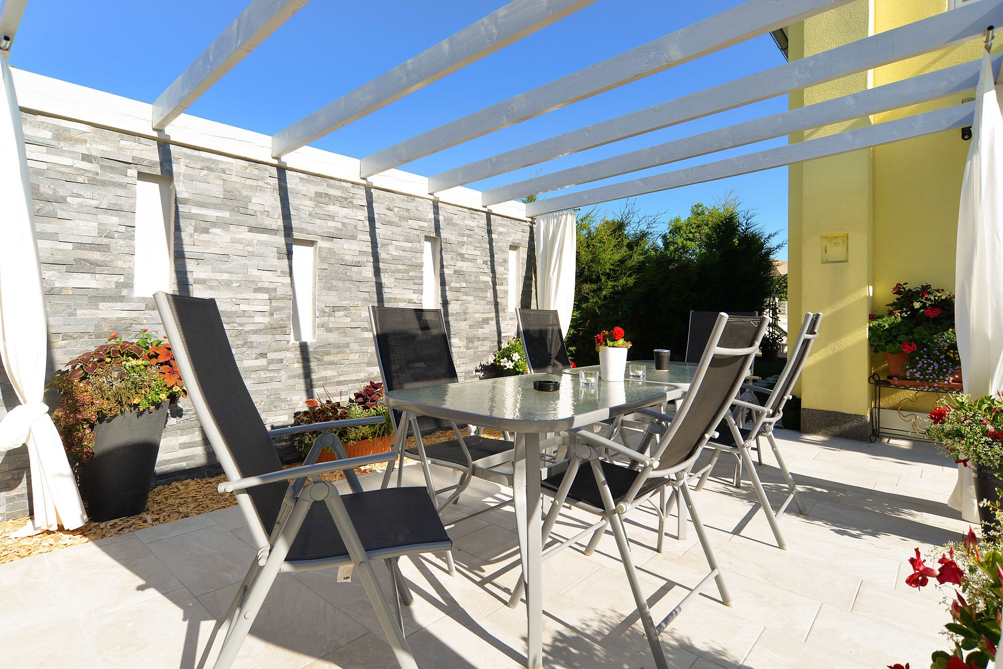 Une terrasse extérieure avec une table en verre et des chaises en métal, entourée de plantes en pot et d’un mur en pierre.