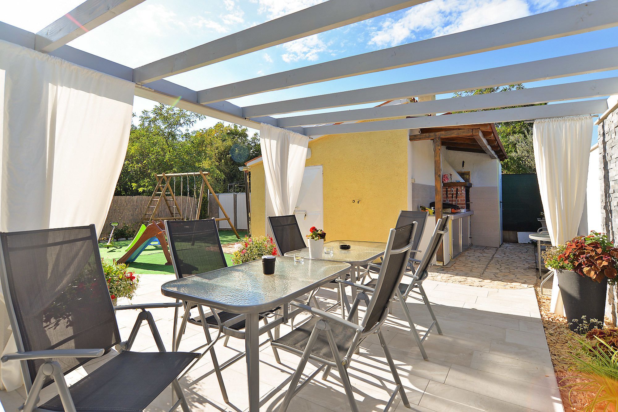 Un patio exterior con mesa y sillas, un columpio y una pared amarilla al fondo.