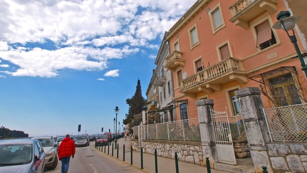 A street with parked cars, a person in a red jacket walking, and a building with balconies and a stone wall. - Apartment 1050 (Rovinj)