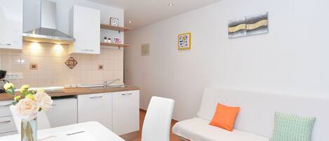 A modern kitchen with white cabinets, a stainless steel range hood, and a wooden countertop. There is a dining table with a vase of flowers and a chair. A white sofa with orange and green pillows is against the wall.