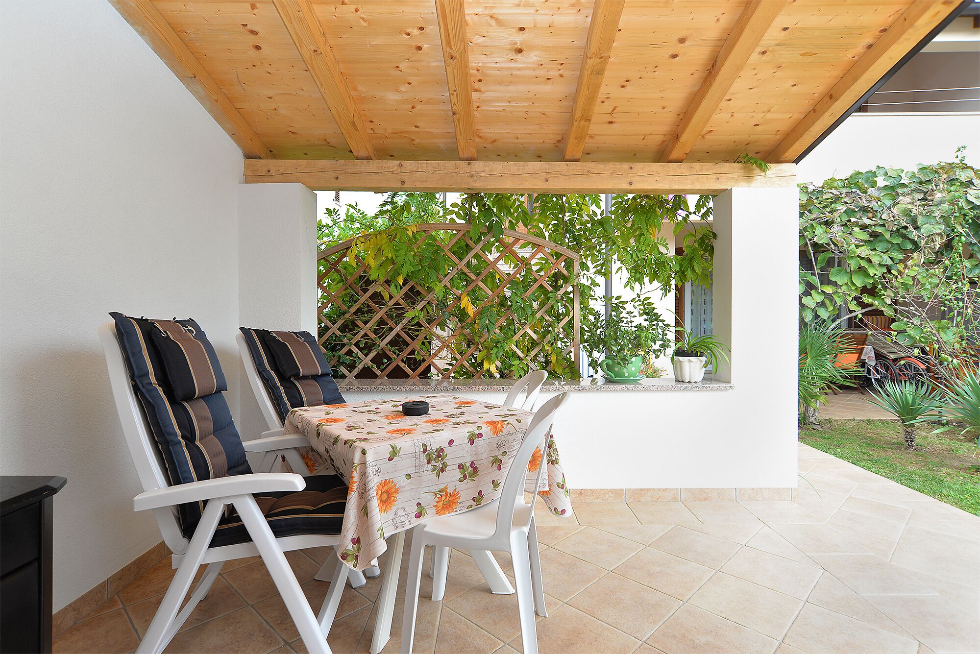 A covered patio with a table and chairs, a wooden ceiling, and a view of a garden with plants and a trellis.