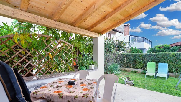 Eine Terrasss mit Tisch, bedeckt mit einer blumigen Tischdecke, weißen Stühlen und einer hölzernen Pergola darüber.