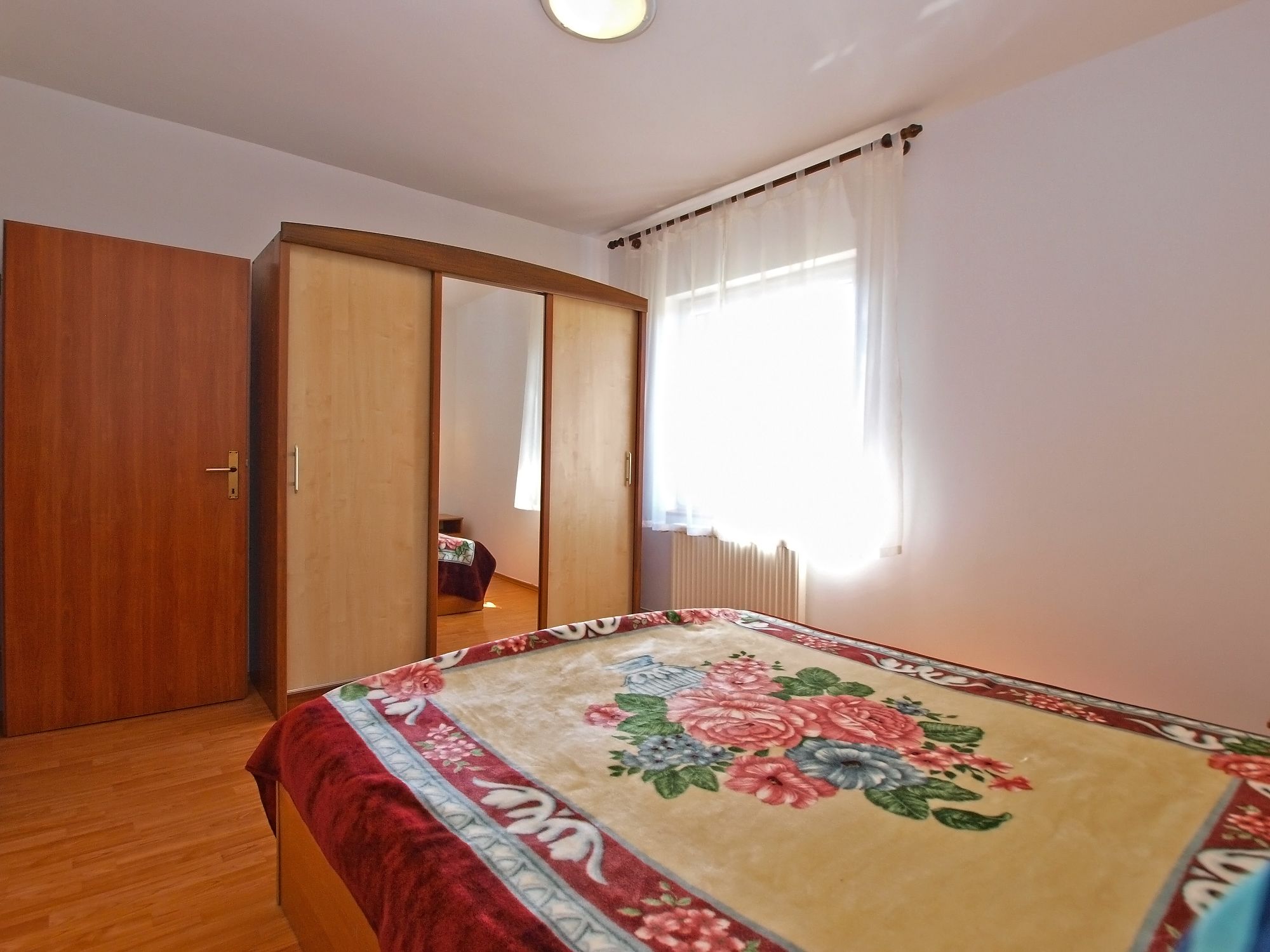 Un dormitorio con un armario de madera, una cama con colcha floral y una ventana con cortinas de gasa.