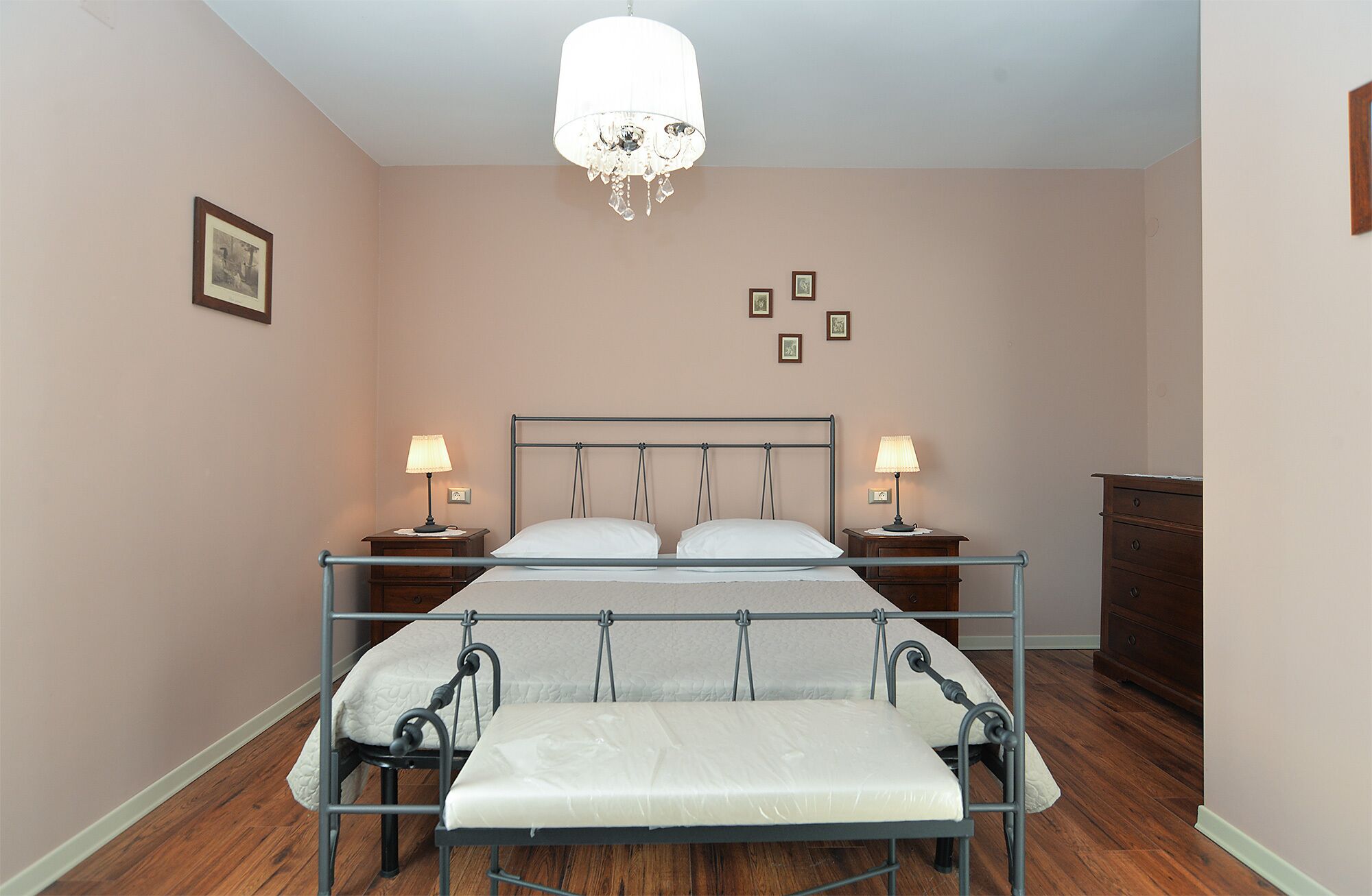 A bedroom with a metal bed frame, two bedside tables with lamps, a wooden dresser, and a chandelier.