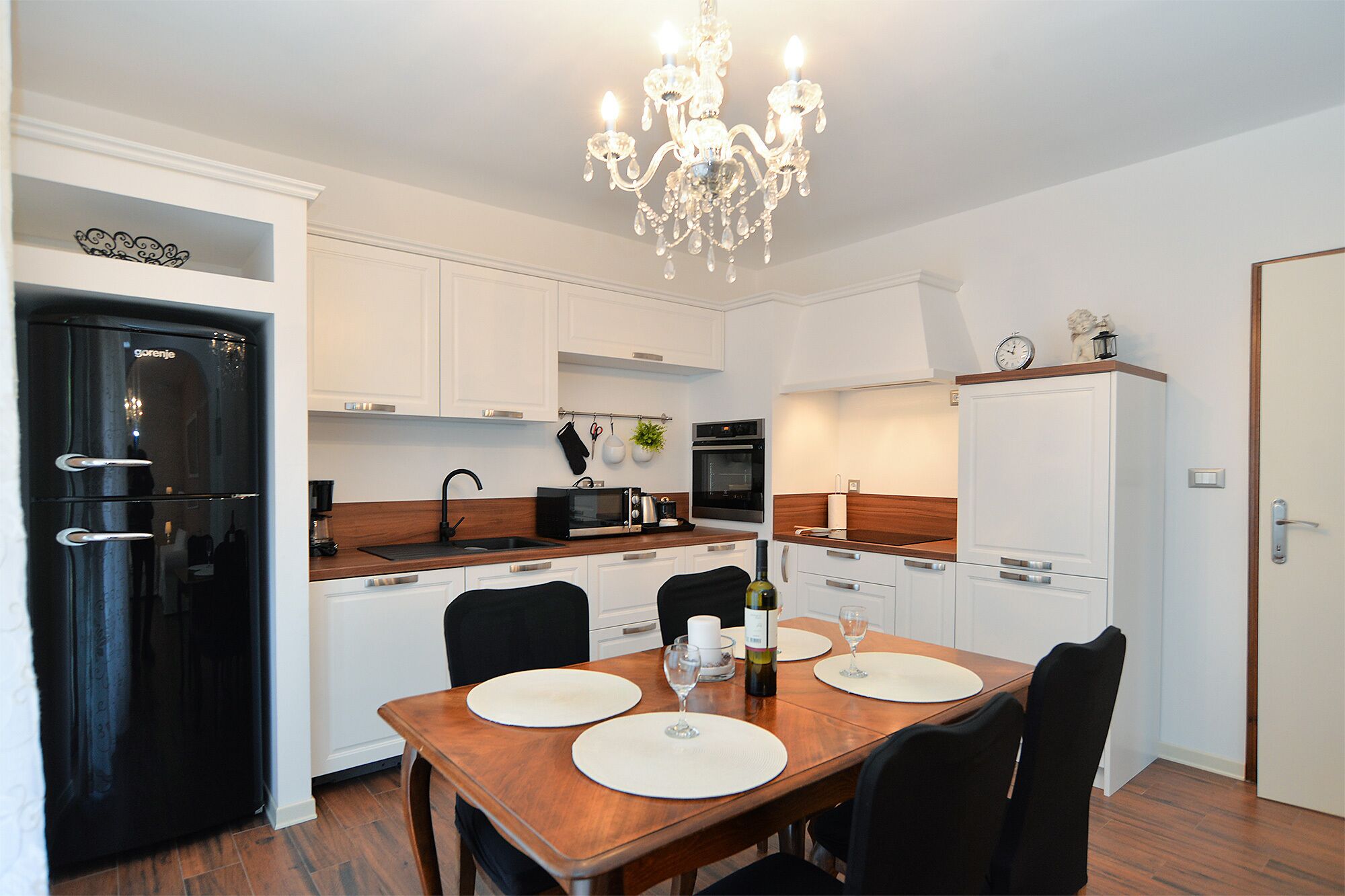 A modern kitchen with white cabinets, a black refrigerator, a wooden dining table with white placemats, and a chandelier.