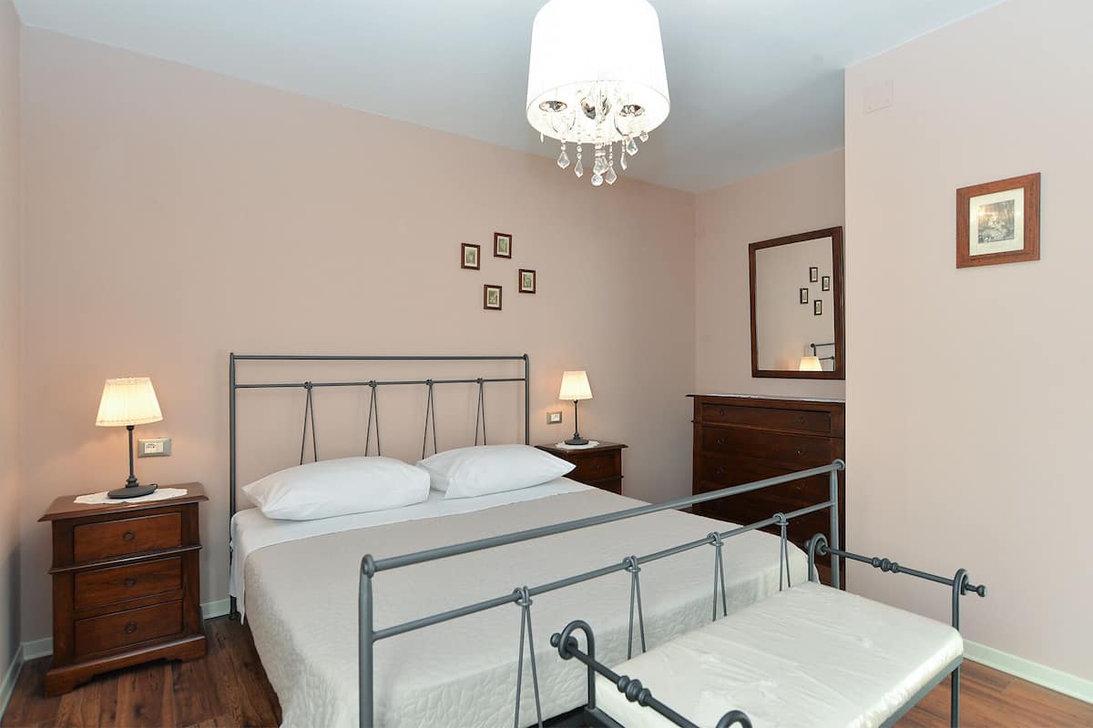 Ein Schlafzimmer mit einem Himmelbett, zwei Nachttischen mit Lampen, einer Holzkommode, einem Spiegel und einer Deckenleuchte.